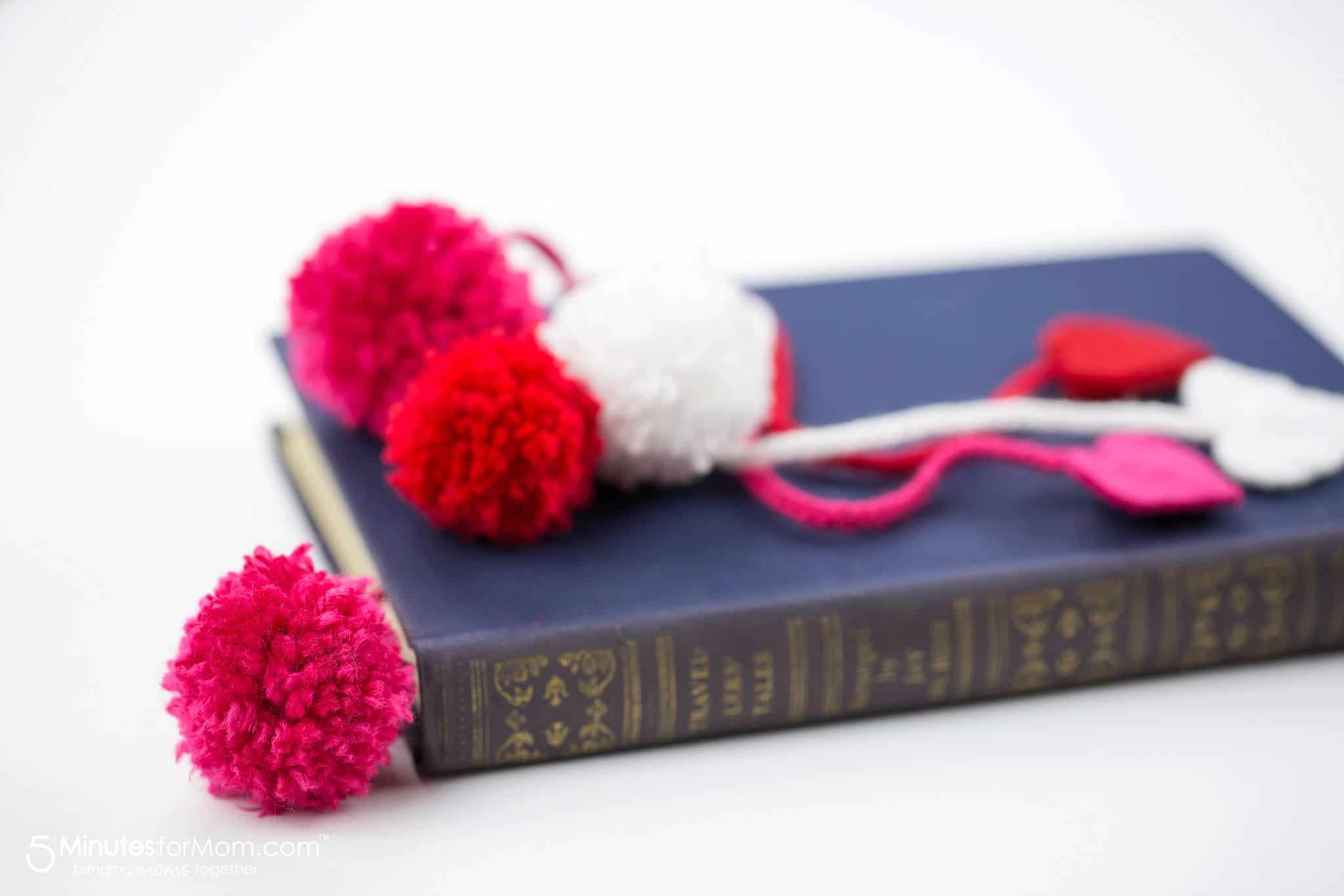Sideview photo of a navy blue hard cover book with a pink pom pom bookmark inside of it. Resting on top of the book are white, red and pink pom pom bookmarks with felt hearts.