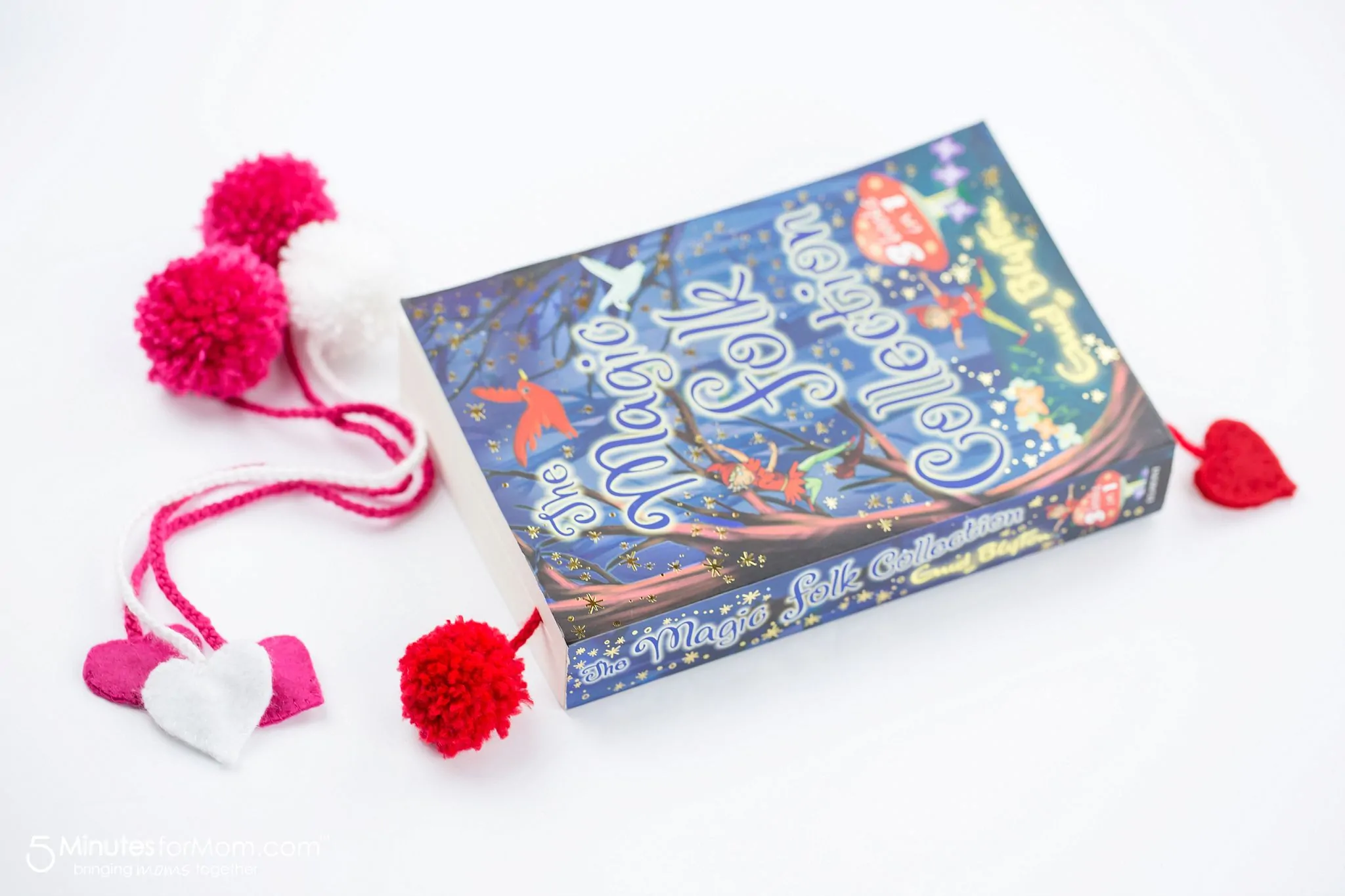 Three DIY pom pom bookmarks sitting next to a book with a page marked by a red Yarn Pom Pom Bookmark.