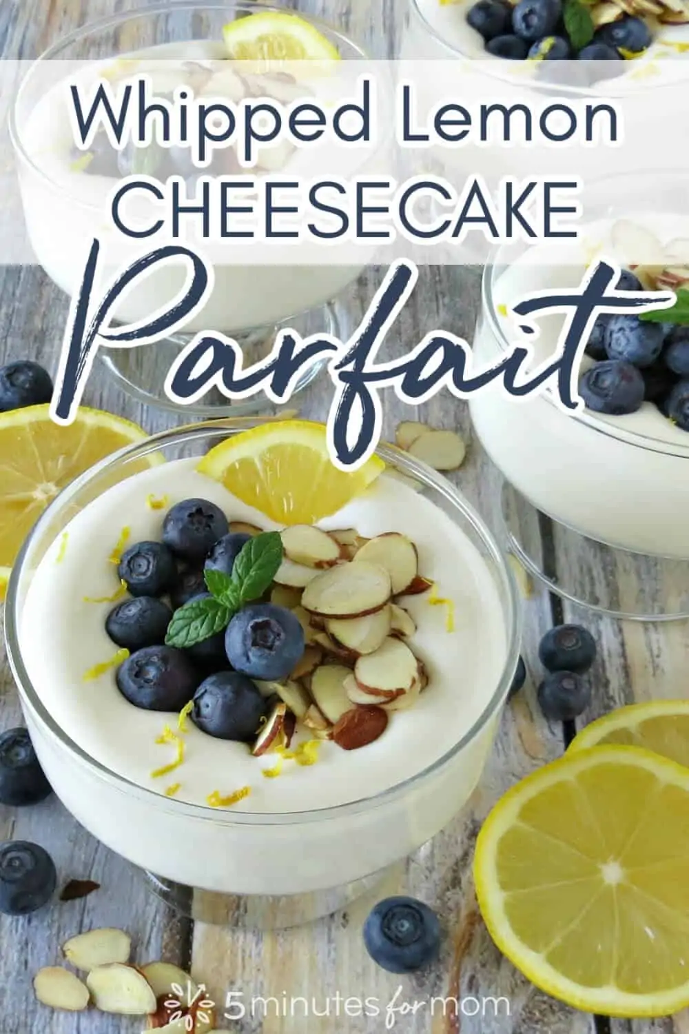 Dessert dish filled with Whipped Lemon Cheesecake Parfait and topped with blueberries, almond slices and a lemon slice. The photo also includes a few other servings of the dessert in the background and some extra lemon slices next to it on the table. Text on image says Whipped Lemon Cheesecake Parfait.