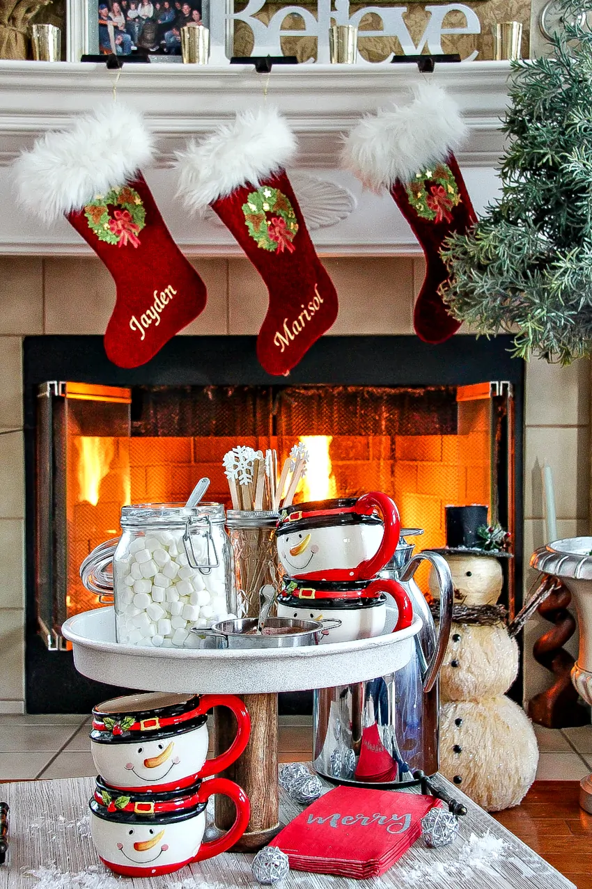 Christmas stockings hanging by a fireplace with a hot chocolate stand and snowman mugs on display.