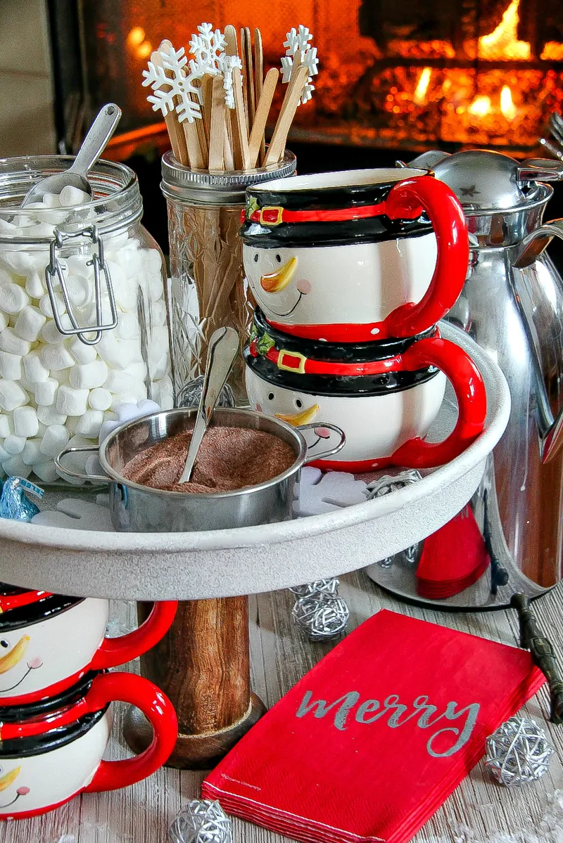 Christmas hot chocolate station with mugs, marshmallows, mixing sticks, and hot chocolate mix sitting on a table in front of a roaring fire in a fireplace.