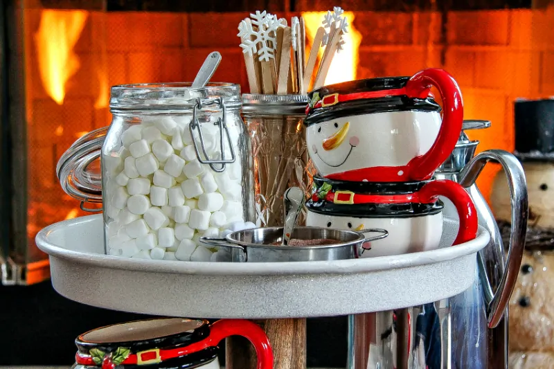 Homemade hot chocolate station with marshmallows, snowman mugs and a fireplace in the background.