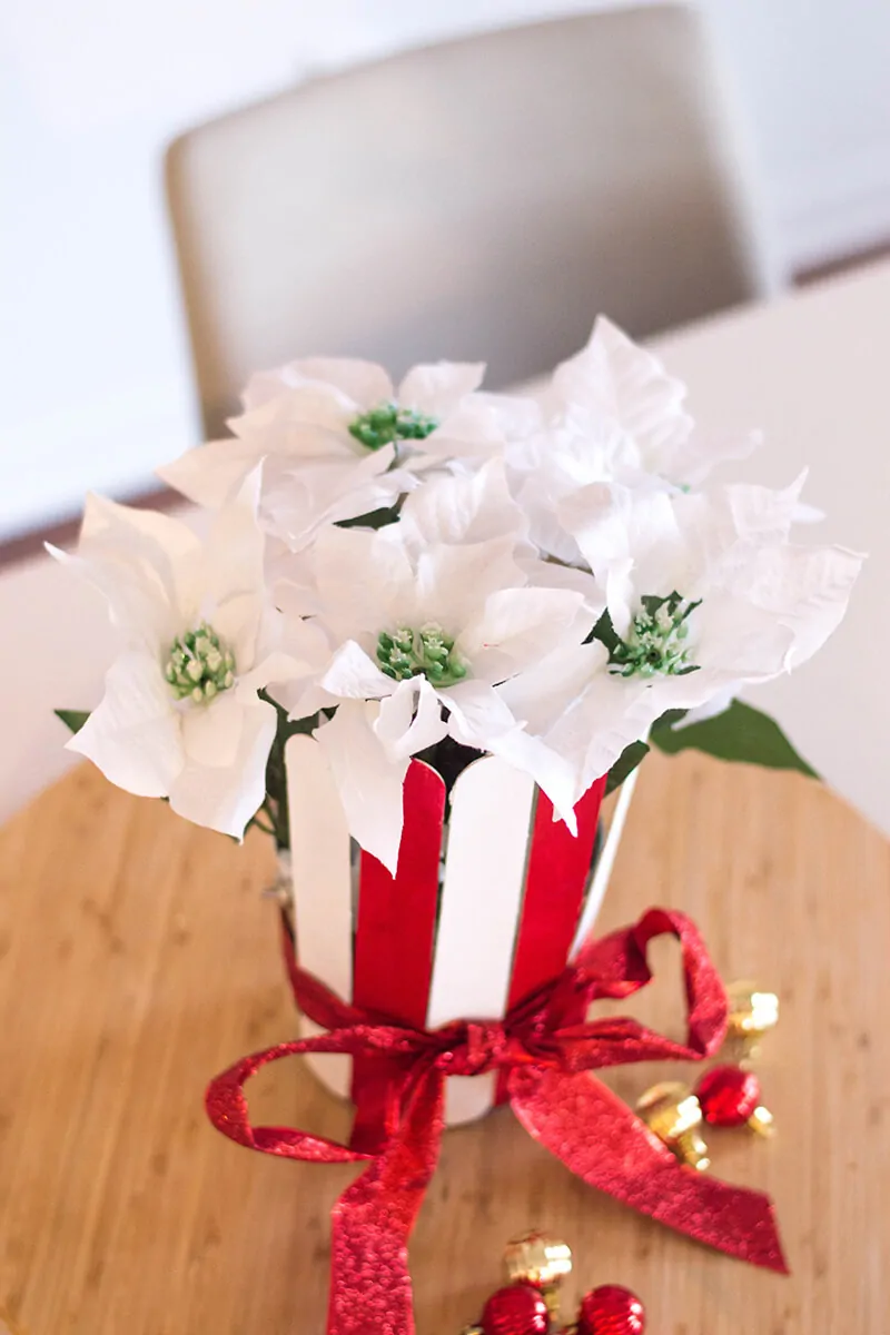 Vertical overhead photo of a white poinsettia in a DIY plant pot made with red and white extra-large popsicle sticks. It has a red ribbon tied in a bow around it and red and gold ornaments on the table next to the pot.