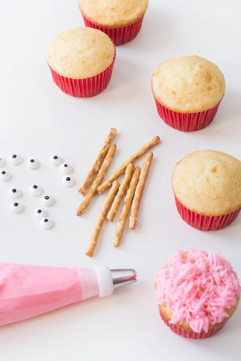 Overhead vertical photo of plain cupcakes, pretzels, pink frosting bag and one pink frosted cupcake.