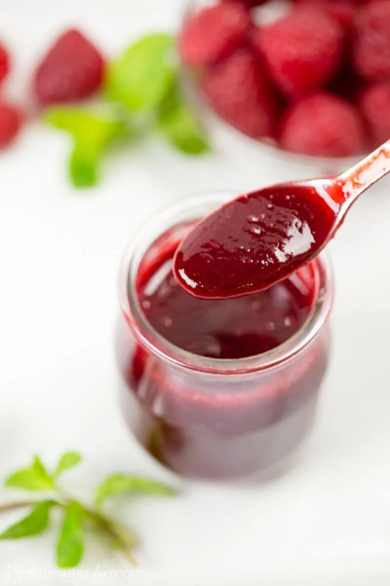 Close up view of a spoonful of raspberry coulis above a small jar of the raspberry sauce.