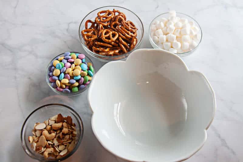 spring snack mix ingredients in clear bowls, and white bowl on white marble background