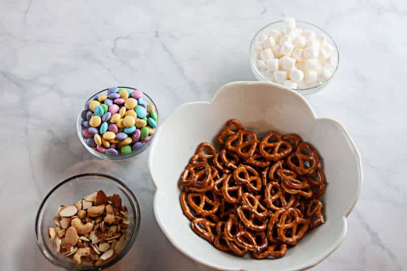 spring snack mix ingredients in clear bowls, and white bowl on white marble background