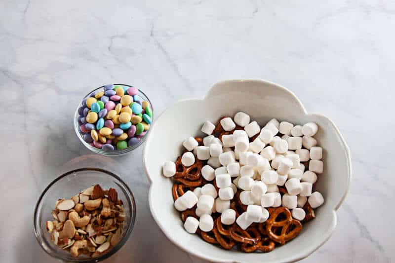 spring snack mix ingredients in clear bowls, and white bowl on white marble background