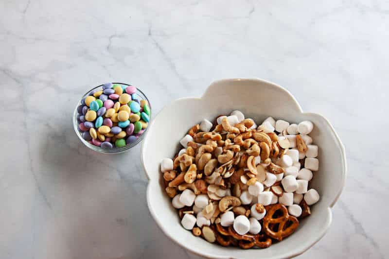 spring snack mix ingredients in clear bowls, and white bowl on white marble background