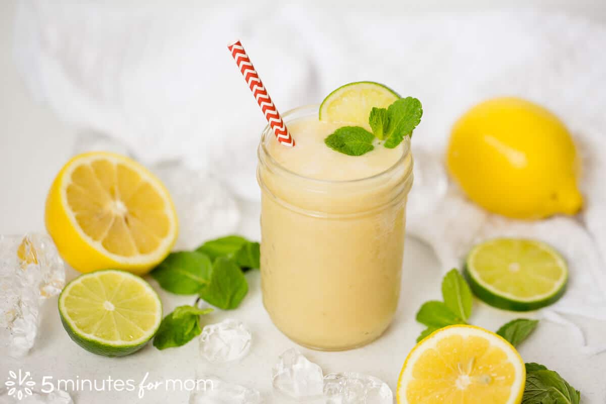 Lemon Iced Tea Smoothie served in a mason jar with a slice of lime and mint garlic, resting on a white table surrounded by sliced lemons and limes and mint leaves.