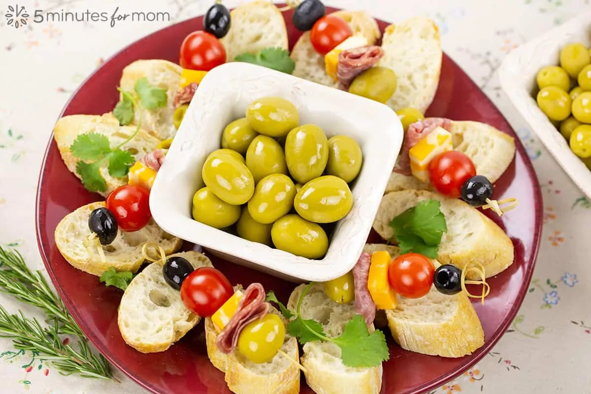 A platter filled with sliced baguette and skewers with European olives, cheese, tomatoes, and deli meat.