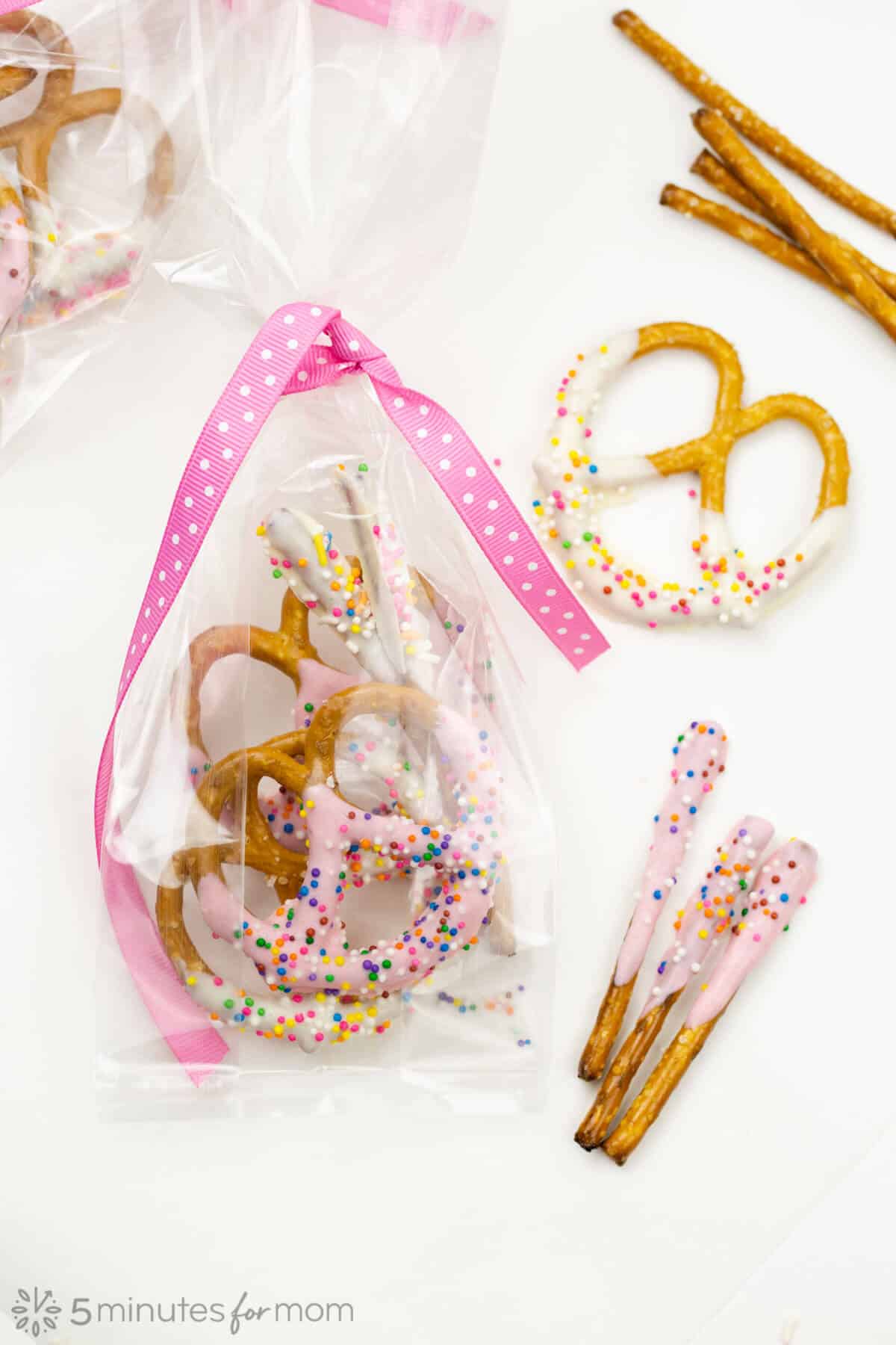 wrapped pink chocolate covered pretzels sitting next to a decorated white chocolate covered pretzel twist and sprinkled pink pretzel sticks.