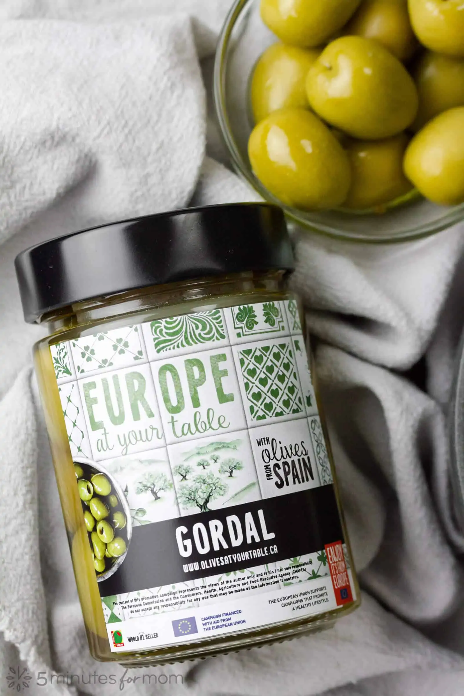 a jar of Gordal olives on a grey linen cloth with a small bowl of large green olives