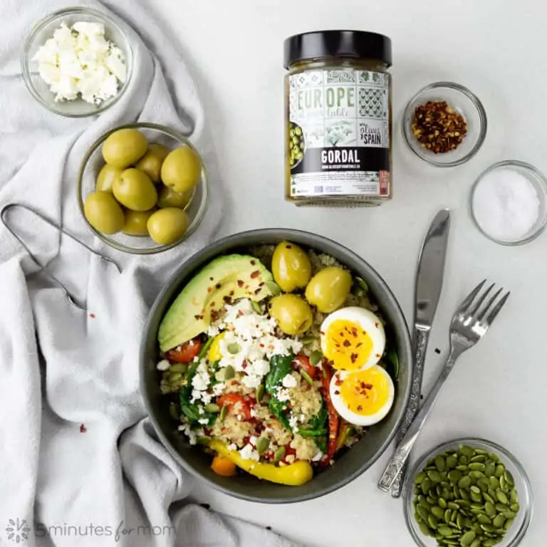 breakfast bowl with quinoa, sautÃ©ed vegetables, avocado slices, soft boiled egg, and three large green olives on a grey table with a jar of Gordal olives, and small bowls of pumpkin seeds, green olives, white cheese, salt, and red pepper flakes