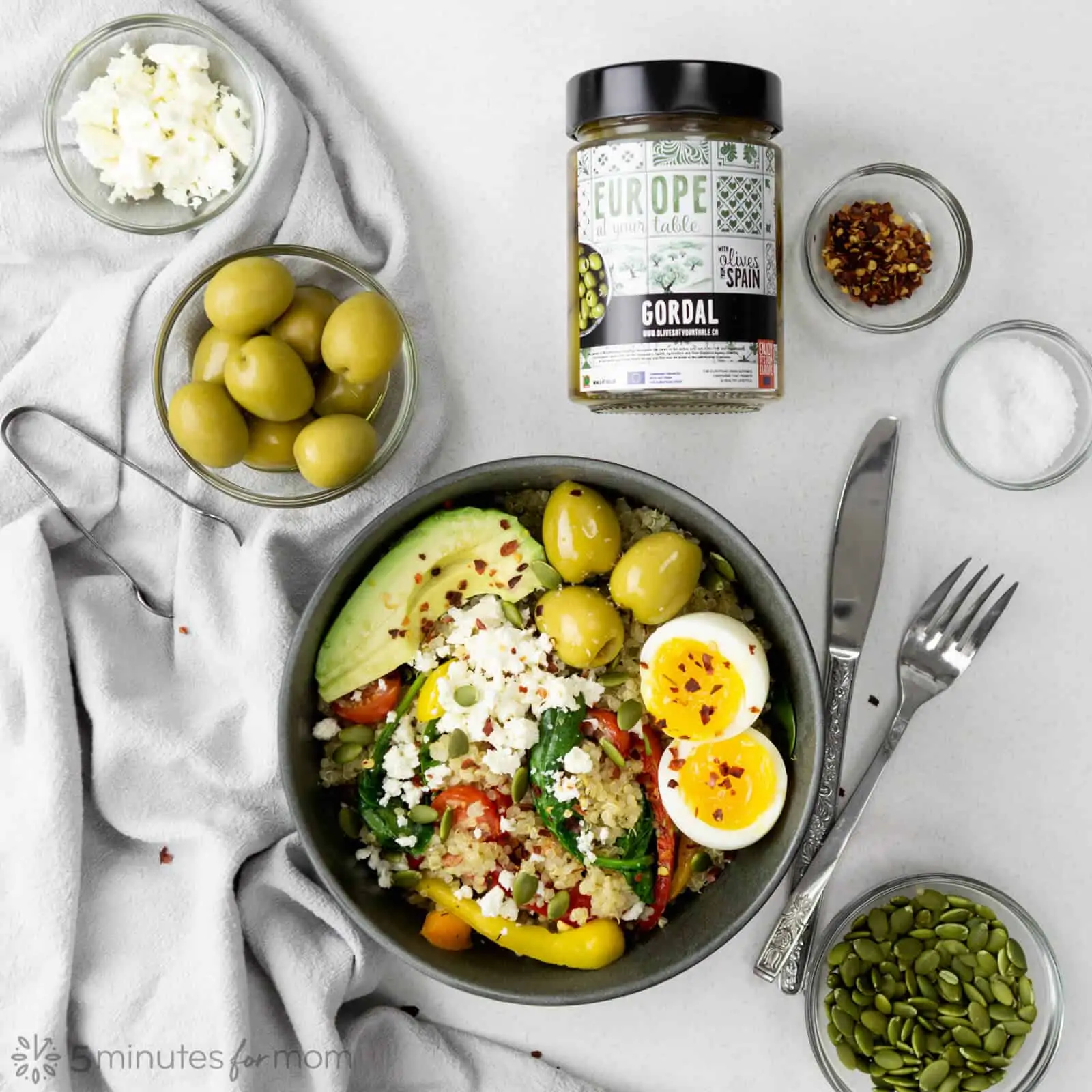 breakfast bowl with quinoa, sauteed vegetables, avocado slices, soft boiled egg, and three large green olives on a grey table with a jar of Gordal olives, and small bowls of pumpkin seeds, green olives, white cheese, salt, and red pepper flakes