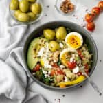 a healthy breakfast bowl with quinoa, sautÃ©ed vegetables, avocado slices, soft boiled egg, and three large green olives, topped with crumbled cheese, pumpkin seeds, and red pepper flakes