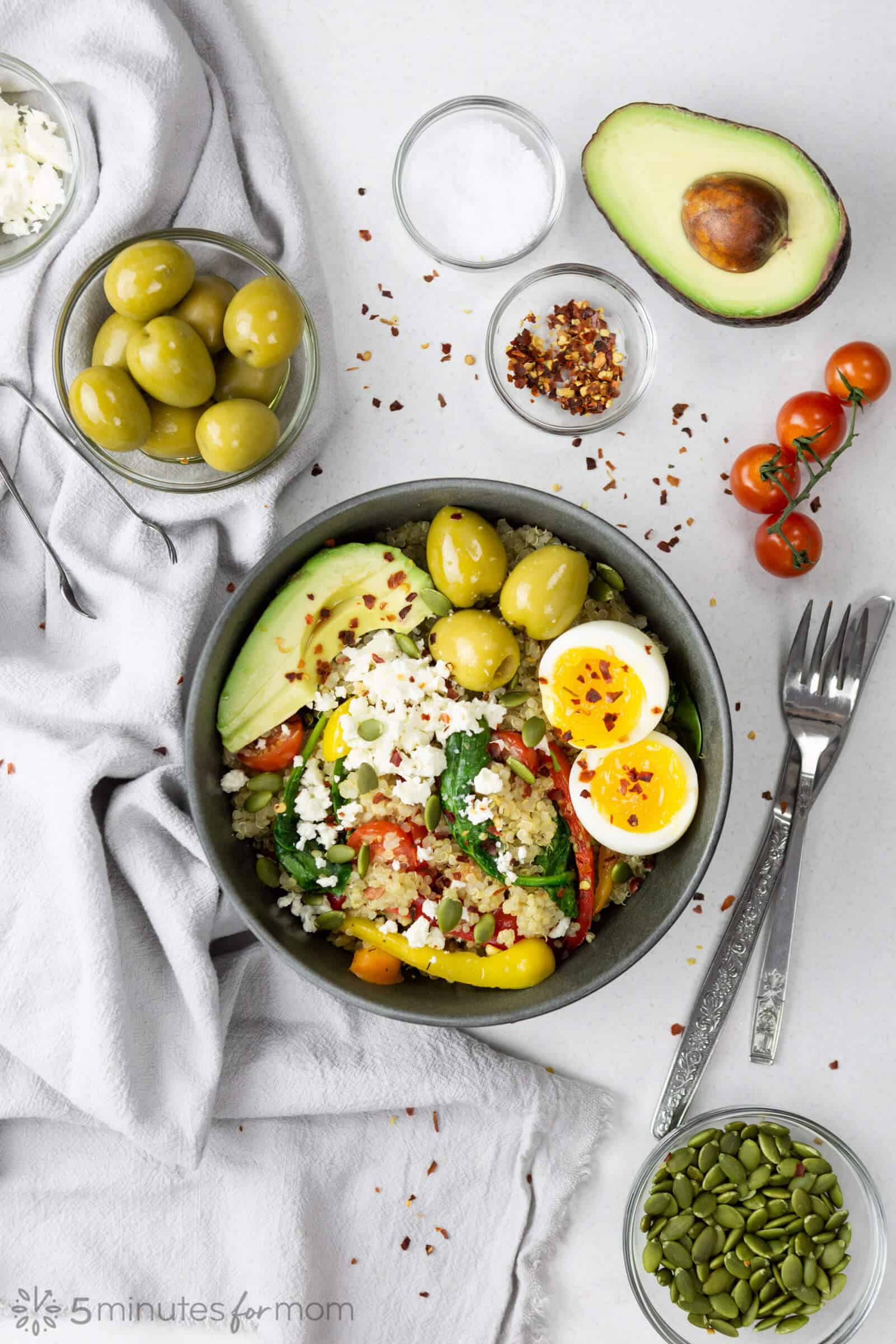 a spread of food on grey table that includes a bowl with quinoa, sauteed vegetables, avocado slices, soft boiled egg, and three large green olives